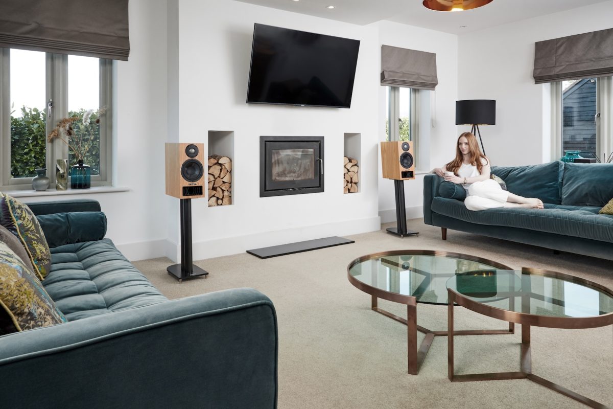 PMC twenty5.22i Standmount Speakers Lifestyle image of woman lying on green velvet sofa with tv on wall and wood burner