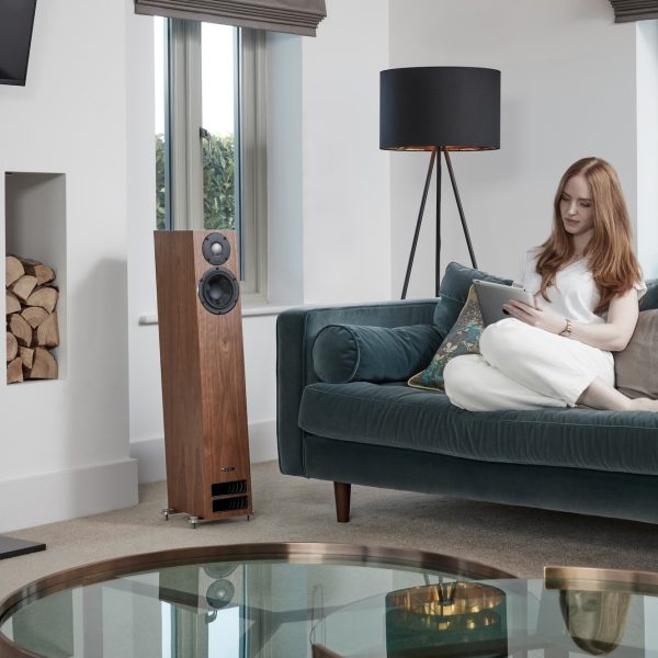 PMC twenty5.23i Floorstanding Speakers lifestyle image with woman on green velvet couch, lamp in corner and glass coffee table in foreground