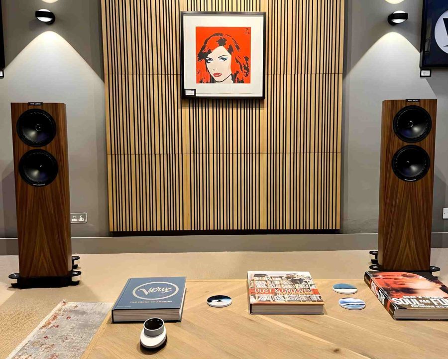 Fyne Audio f702sp floorstanding speaker in natural walnut finish straight view in demonstration room with artnovion siena treatment on wall with debbie harry pop art picture and coffee table with verve records book